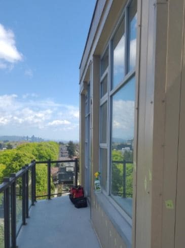 A narrow upper-floor balcony with a smooth grey coated deck and a glass railing, looking out over trees to the city