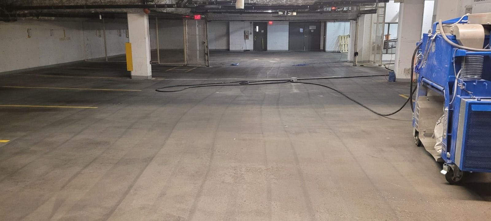 An underground parkade deck with parallel pass marks from shot-blasting, the blue dust collector at the right